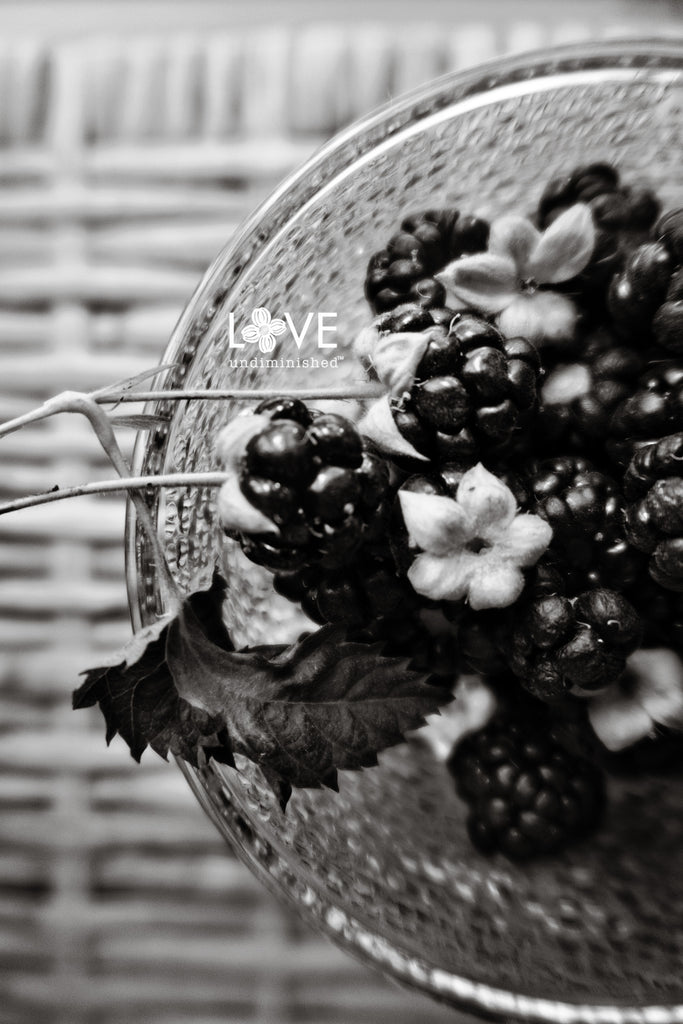 Black and white photograph of hand-picked fresh, plump blackberries from the garden, in a clear glass bowl on a wicker table.